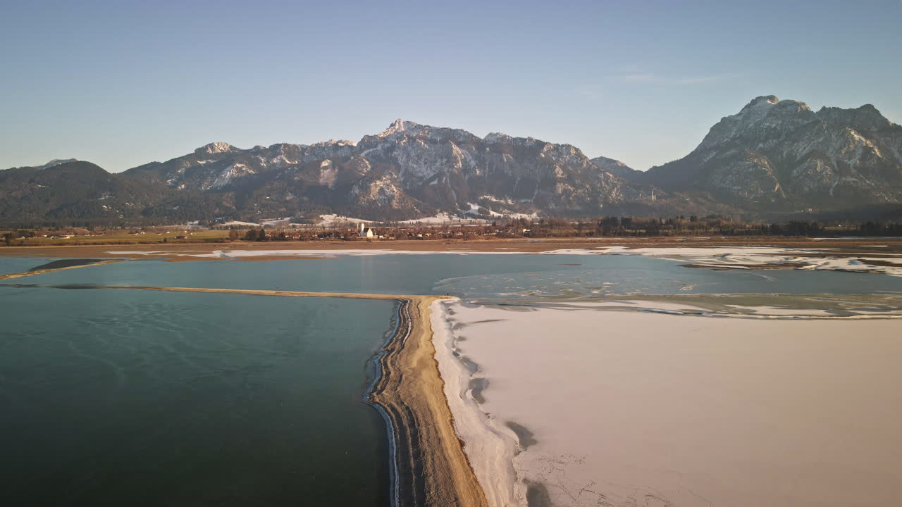 Camera descends, revealing a striking transition from frozen white surfaces to clear water and snow-lined sandbars with mountain backdrops