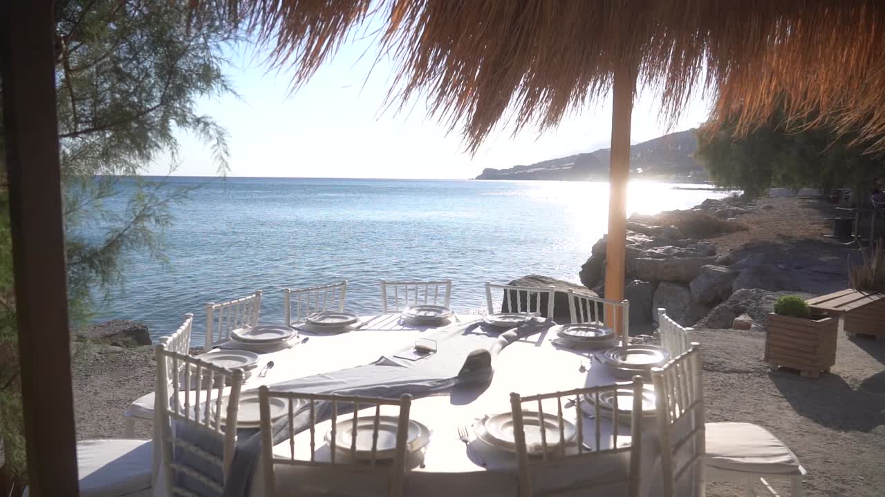 Filmische Zeitlupenaufnahme, die an einem Strohschirm am Strand vorbeiführt und einen dekorierten runden Tisch enthüllt, während Sonnenlicht auf dem ruhigen Meer reflektiert wird, wodurch eine warme und malerische Atmosphäre entsteht