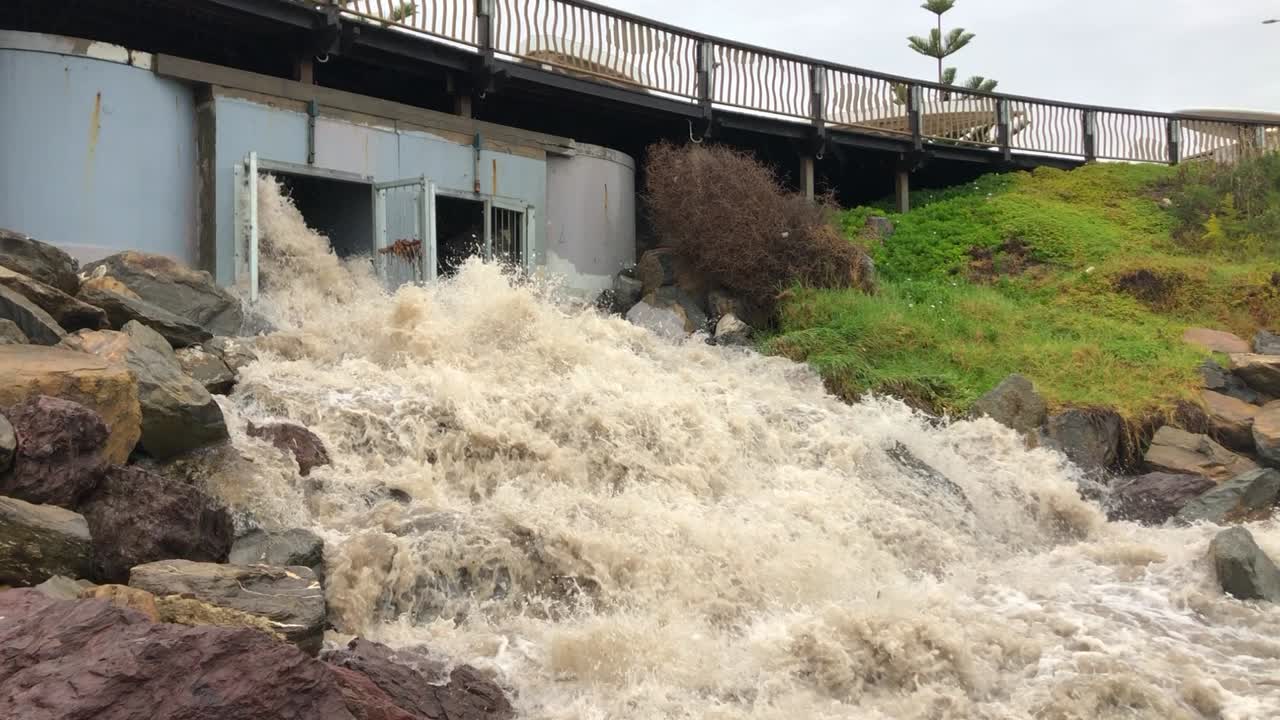 El agua de la inundación surge de una salida de drenaje hacia el océano, lo que ilustra las consecuencias de las fuertes lluvias y las posibles consecuencias medioambientales.