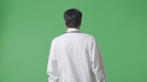병원 에서 녹색 스크린 배경 에 걸어가는 스테토스코프 를 들고 있는 아시아 남자 의사 의 뒷면