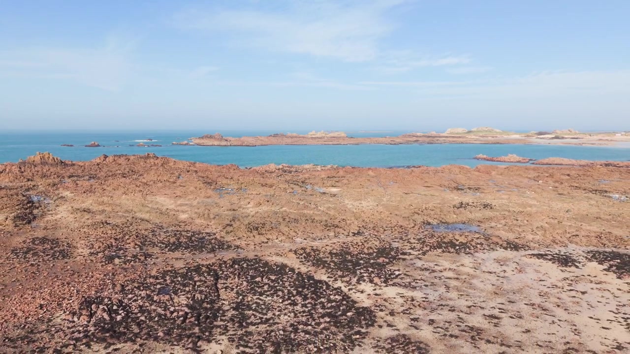 Reveal of rocky then sandy foreshore of bay with calm sea and gentle waves in late afternoon sun against granite outcrops and low headland with blue sky and views to the horizon