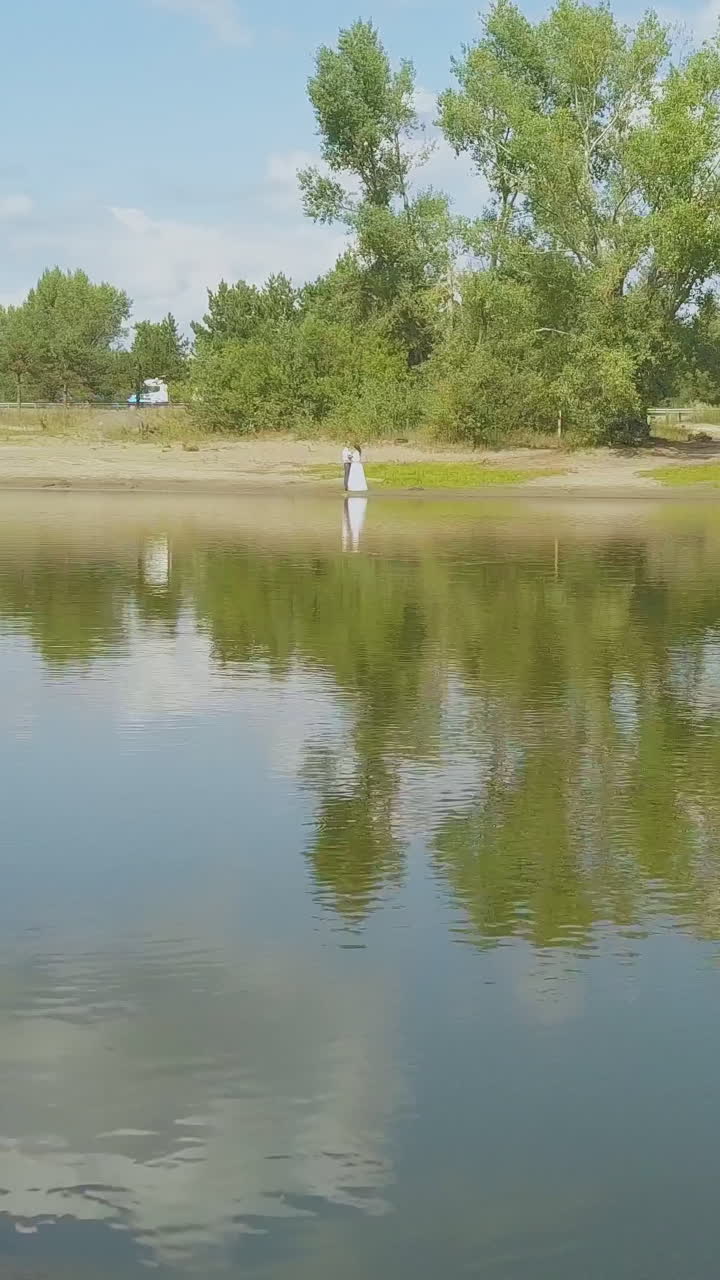 distant newlywed couple rests on bank of wide lake against road and green park on autumn day upper view