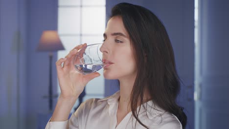 cinematic shot of young smiling woman is drinking fresh pure water from a transparent glass in living room at home. she maintains her body healthy and feels well being.