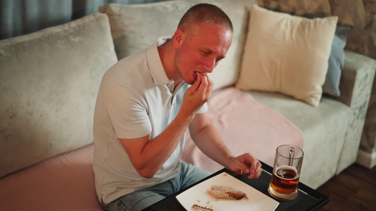 Remote worker seated indoors carefully eating fish to avoid bone from hooking throat, dropping bone pieces on plate, sipping grape wine beside on small table
