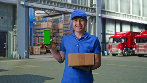 asian male courier in blue uniform smiling and showing green screen smartphone while delivering a carton, outside of logistics distributions warehouse