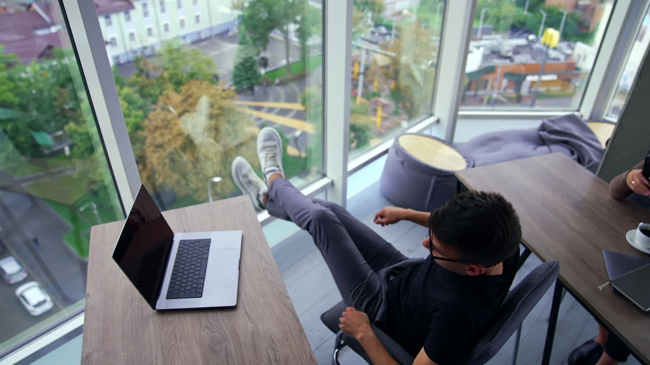 Young man had a break from work and decided to go back to work. Lady at desk behind him speaking on her phone. Office with panoramic windows from high angle view. City from top at backdrop.