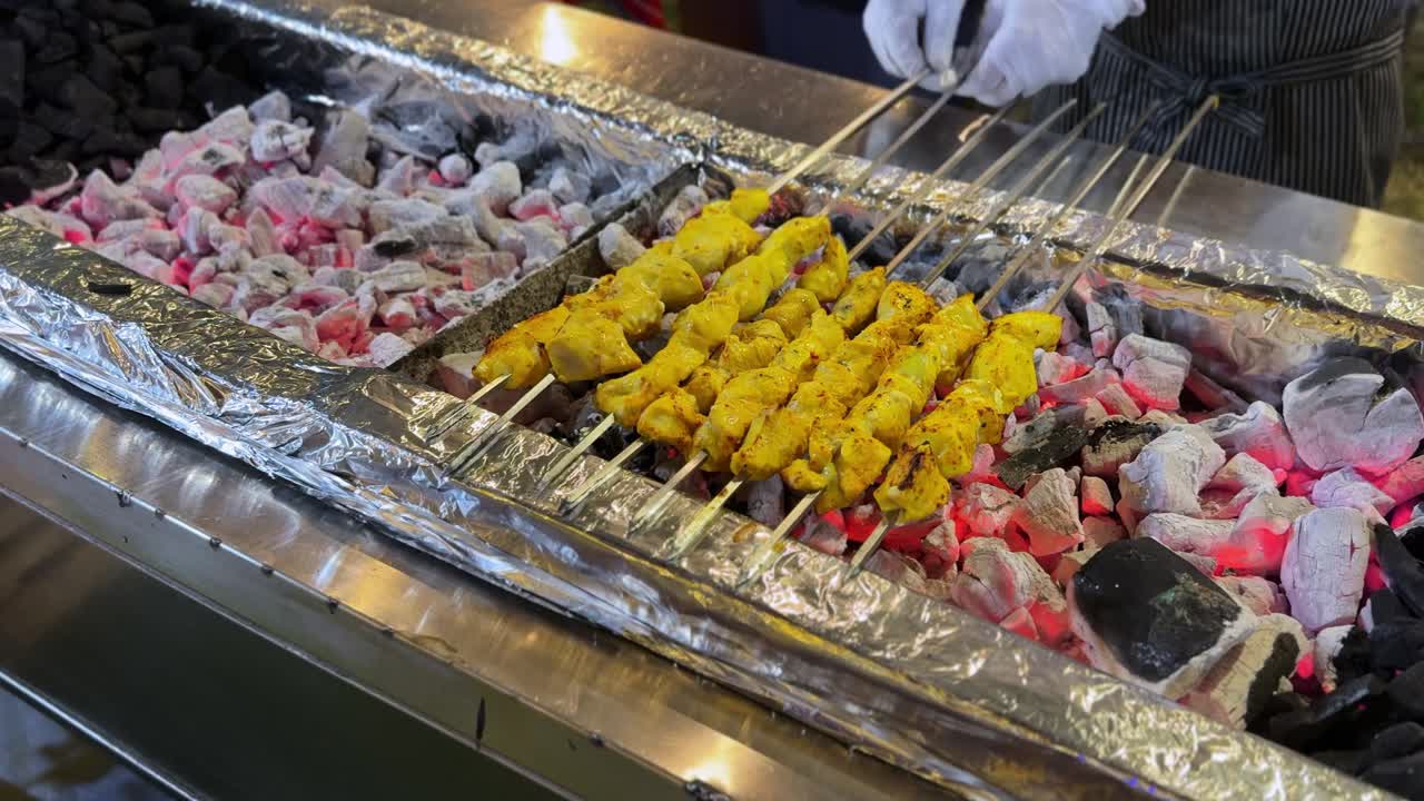 Primo piano di spiedini di kebab di pollo marinato che arrostiscono su braci incandescenti mentre uno chef con i guanti sistema le aste metalliche su una postazione barbecue esterna.