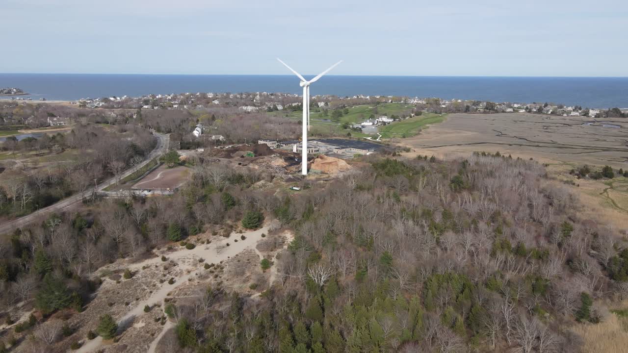 Drone push forward over marshes toward wooded preserve, dog park and wind turbine. Scituate harbor and Atlantic Ocean in the distance.