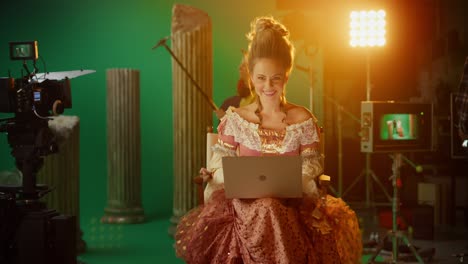 beautiful smiling actress wearing renaissance dress, sitting on a chair using laptop computer with green screen in the background. on film studio period costume drama film set