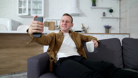 young man taking photo of himself at home. handsome guy sitting on sofa using mobile phone and drinking tea indoors. smiling man enjoying sitting at home during quarantine.