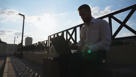 joven empleado trabaja en un cuaderno sentado en un banco en la ciudad. hombre de negocios en ropa formal usando la computadora en la cidade. empresario exitoso escribiendo texto en la computadora portátil al aire libre. concepto de finanzas. dolly shot