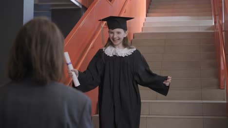 feliz estudiante de preescolar con toga y birrete corriendo por las escaleras, sosteniendo un diploma de graduación y abrazando a la maestra