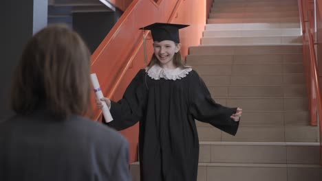 happy preschool female student in cap and gown running down the stairs, holding graduation diploma and hugging teacher