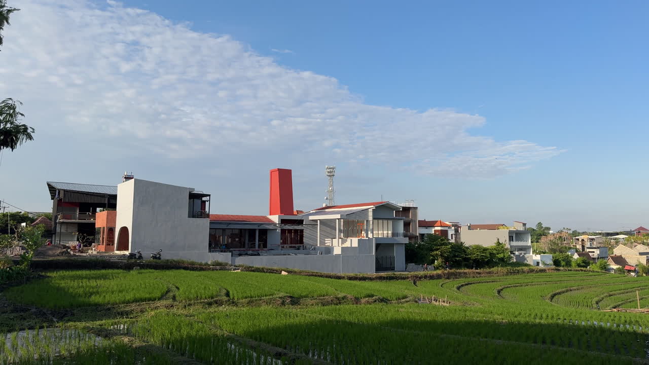 Modern buildings with distinctive architecture stand beside lush green rice fields under a clear sky, showcasing rural-urban contrast.