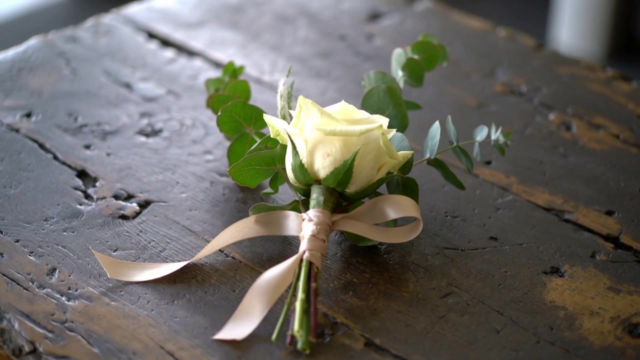 Cámara alejándose para revelar un prendido sobre una mesa desgastada, destacando una rosa blanca, eucalipto y una cinta de satén.