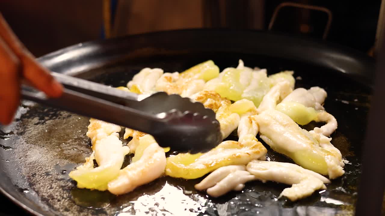 Hand uses tongs to fry squid eggs on a griddle at a Bangkok night market