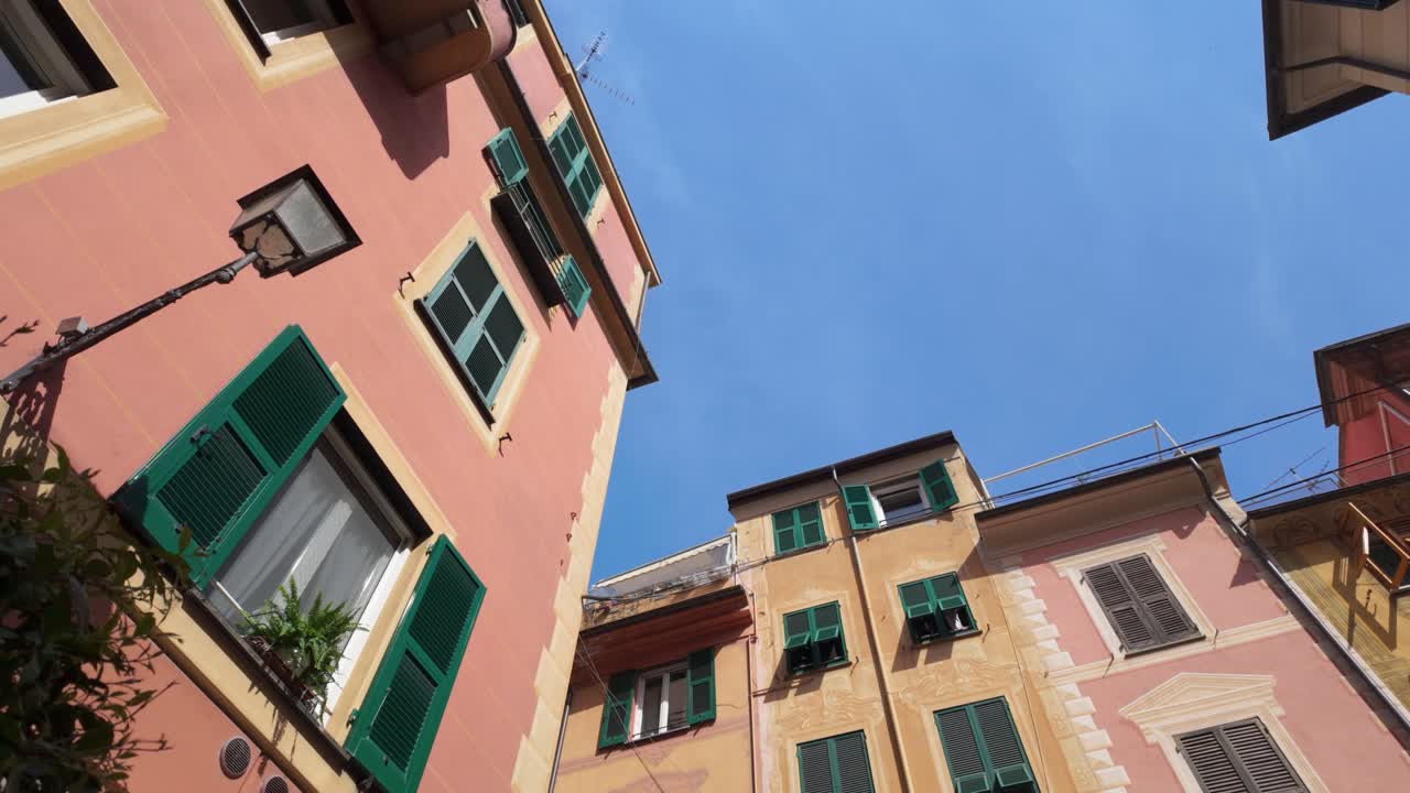 Vista em grande angular de edifícios em tons pastel com persianas verdes em um pátio europeu ensolarado, apresentando arquitetura clássica, cores vibrantes e um céu azul claro; sem pessoas presentes.