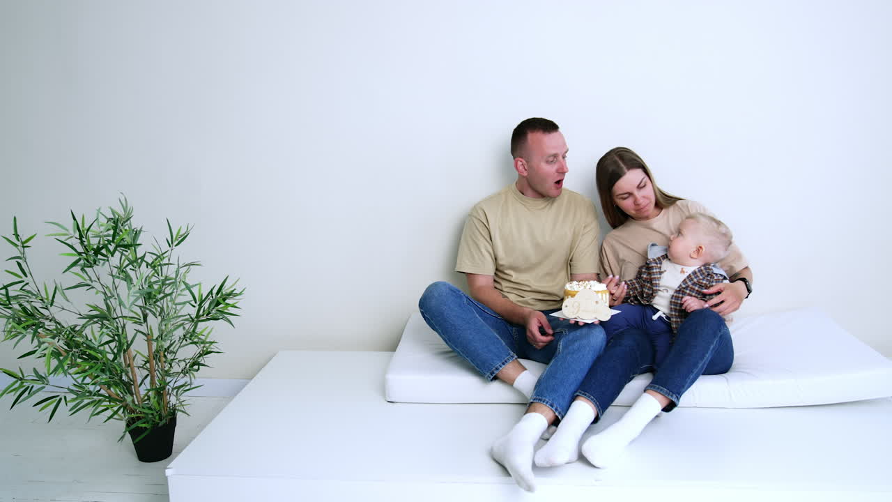 Caucasian parents wearing beige t-shirts and jeans sit at the wall. Baby in mom's hands looks at the cake in dad's hands. White backdrop.