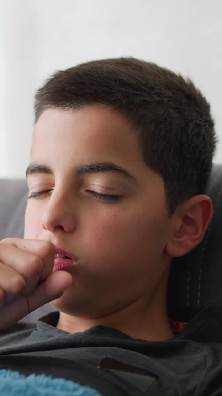 Little kid with brown eyeball looks visibly sick and exhausted as he lies on couch near window coughing and adjusting his position, expressing flu symptoms and discomfort in calm home environment