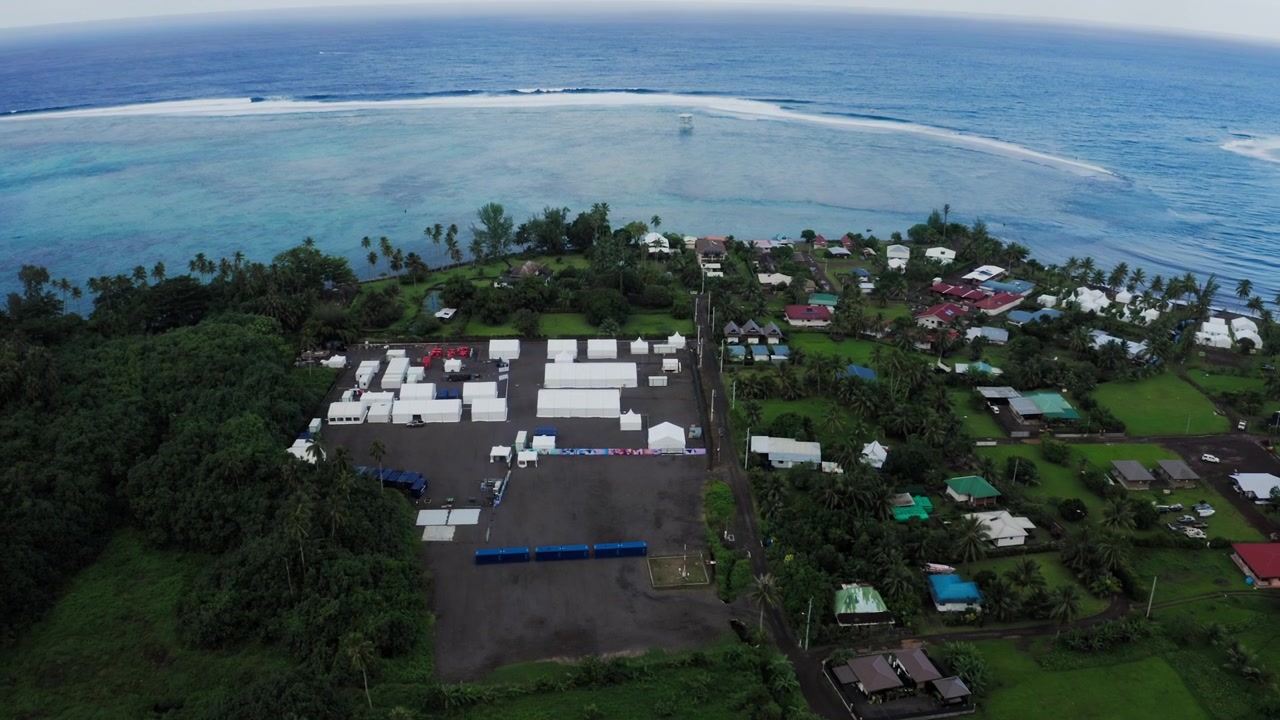 Ampia ripresa aerea con drone su terreni agricoli che confinano con il villaggio di Teahupoo, Tahiti, con campi verdi che si estendono tra la strada costiera e il fianco della montagna.