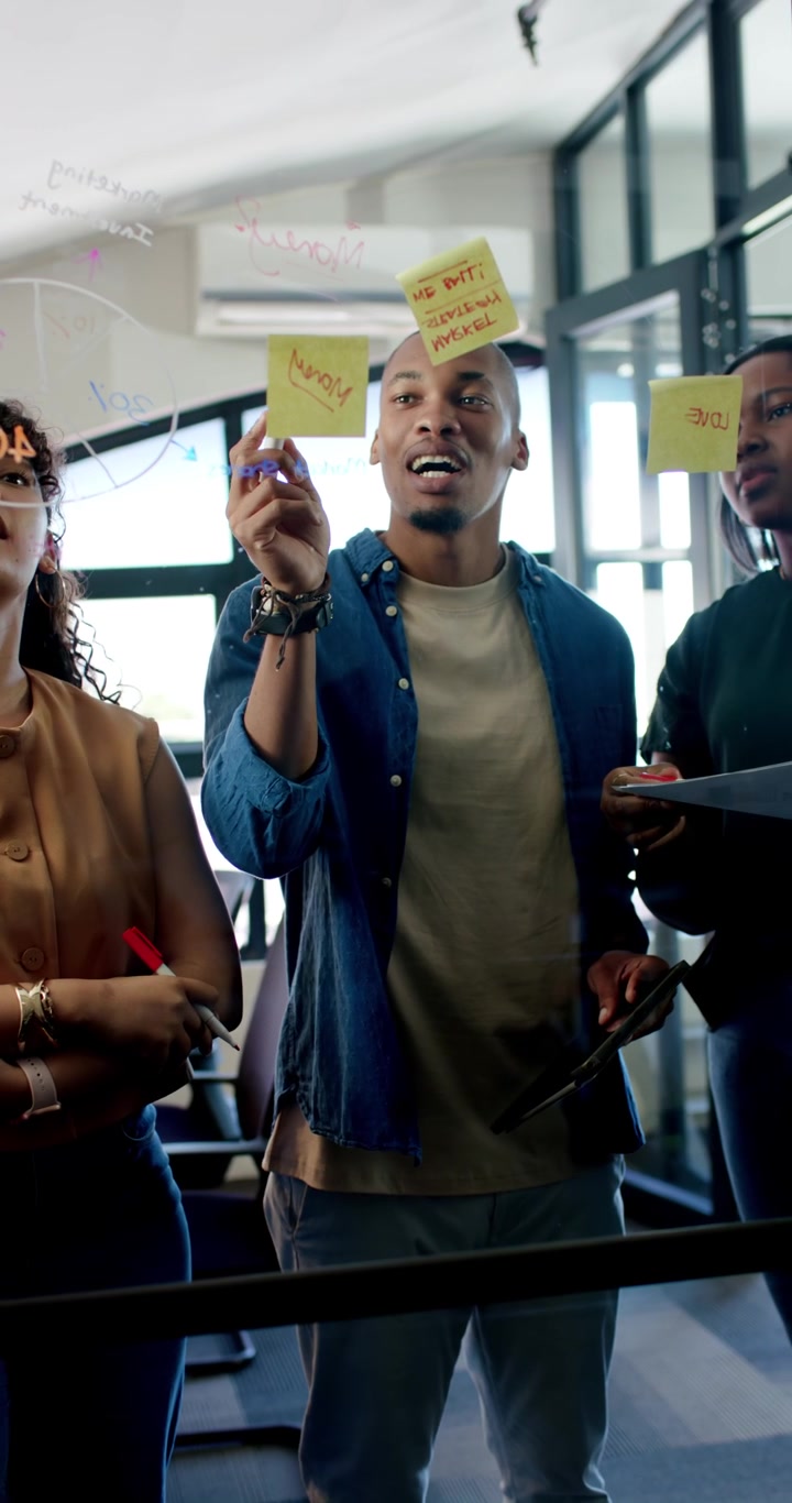 Vertikal video: Divers team skriver på glas med orange tusch, sætter sticky notes op og planlægger.