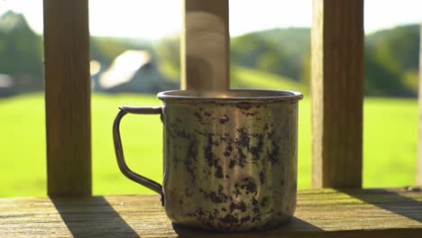 a rustic metal mug steaming in the warm sunlight, overlooking a lush green landscape from a wooden deck
