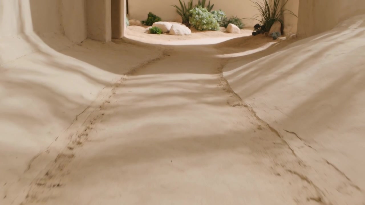 La cámara avanza revelando un camino de arena con dos huellas, márgenes y un orbe pálido en el patio.