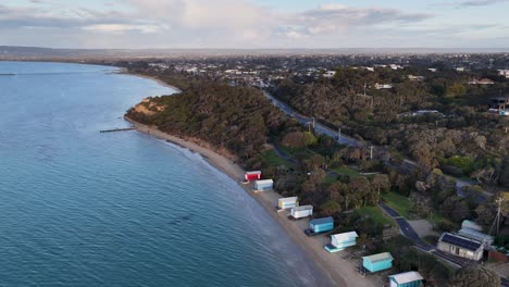 drone glides above shoreline, revealing vibrant bathing boxes, calm sea, and coastal landscape at dusk