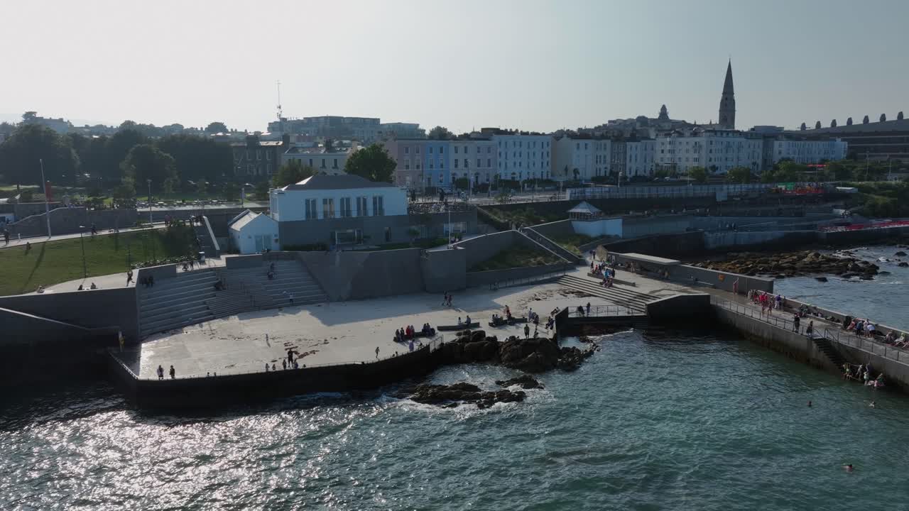 The Baths, Dún Laoghaire, County Dublin, Ireland, September 2024. Drone orbits counter clockwise around the pier with swimmers jumping into the water beside the Roger Casement Statue in sunny weather.