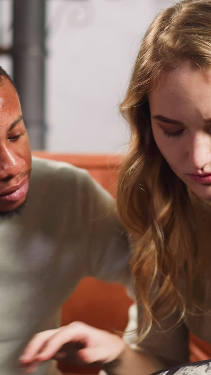 Pretty fair haired woman with African-American husband turns pages of old family photo album sitting on couch in apartment slow motion