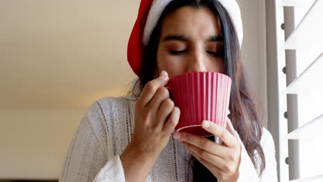 christmas time, indian woman in santa hat enjoying hot drink at home, feeling cozy