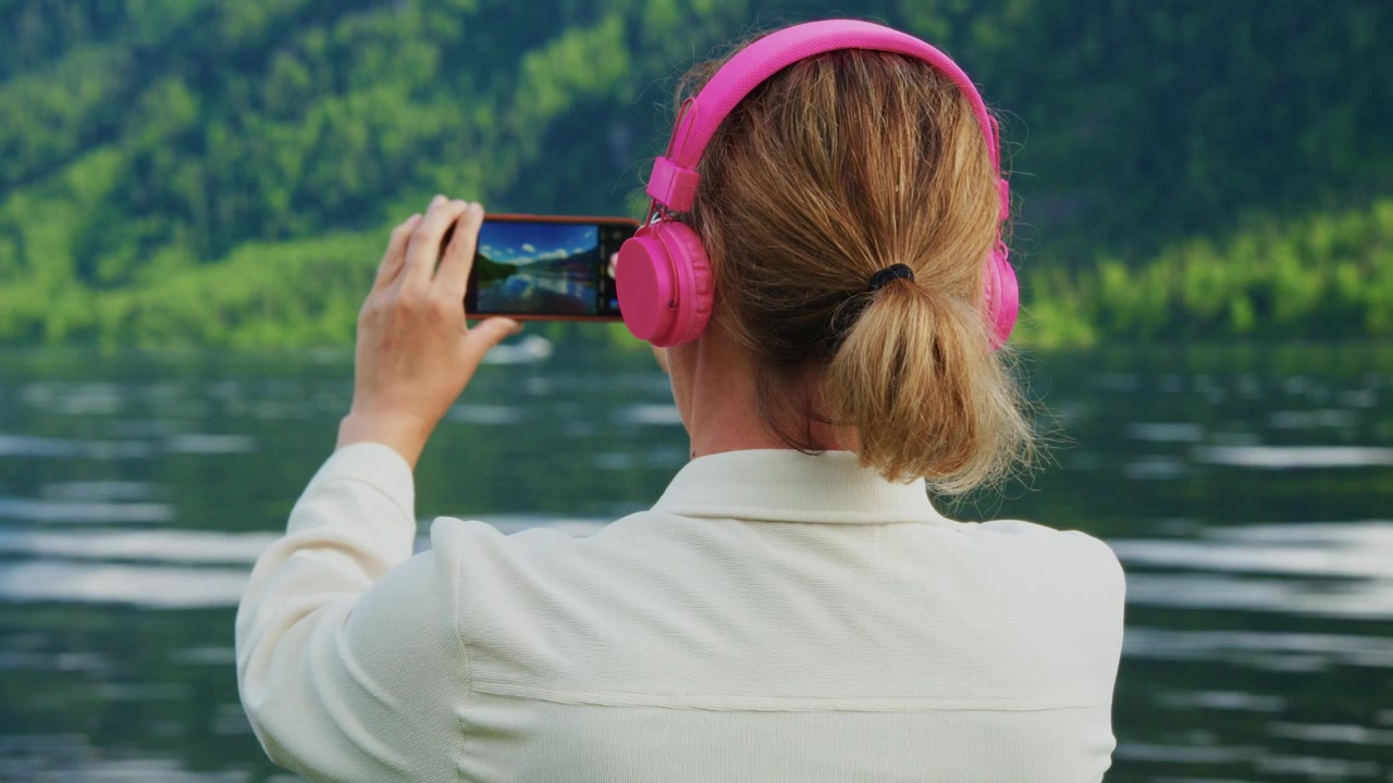 Woman with pink headphones holds phone to capture scenic view. She stands near calm water with lush green forest in background. Her posture shows calm focus as she records tranquil nature