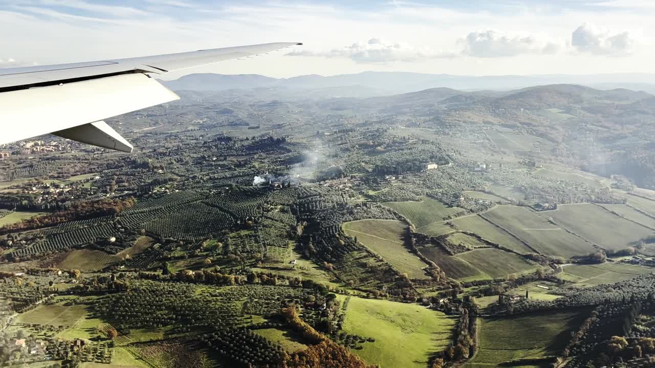 Luftfoto fra et flyvindue over Toscana, Italien, der viser lappetæppe-landbrugsjord, olivenlunde og bølgende bakker med en flyvinge i billedet, klar dagtidsrejse