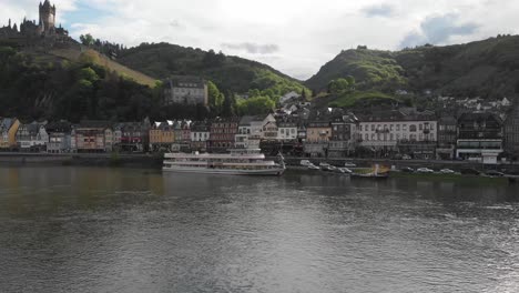 Drone-Despega-Con-Vistas-A-La-Ciudad-De-Cochem-Y-Un-Barco-Fluvial-Junto-A-La-Orilla-Del-Río