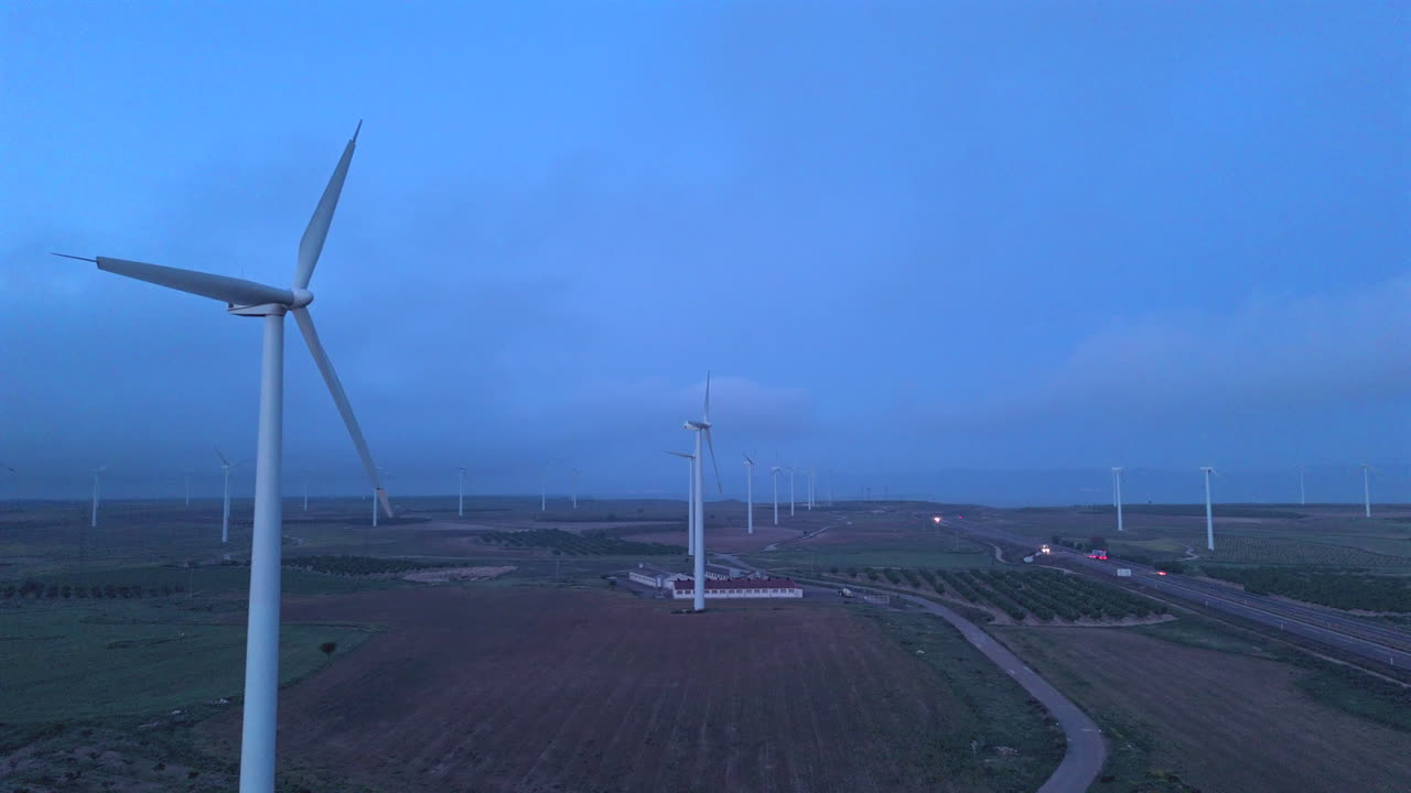 Aerial tracking shot with drone flying backward between wind turbines in a rural wind farm at dawn. Farmland, infrastructure, and highway visible. Smooth cinematic view of sustainable energy