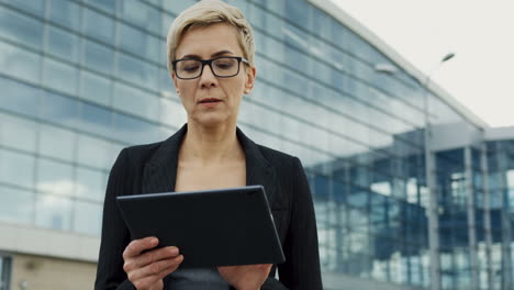 Porträt-Der-Geschäftsfrau-Mit-Brille-Und-Kurzen-Haaren-Auf-Dem-Tablet-Computer-In-Der-Nähe-Eines-Großen-Modernen-Bürogebäudes