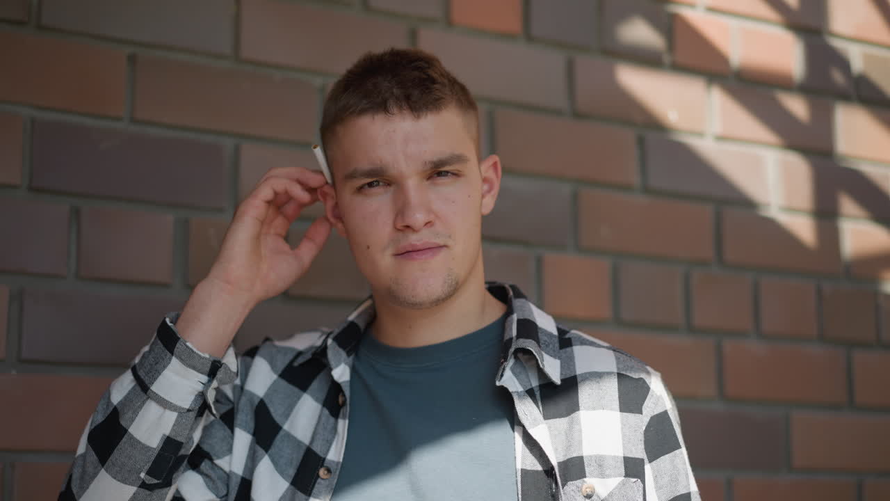 portrait view of young adult in black white plaid shirt on staircase landing returning cigarette to ear while gazing into distance against sunlit brick wall backdrop with metal railing