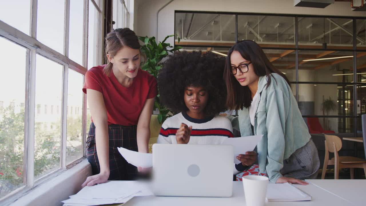 Tre donne millennial che lavorano insieme con un laptop a una scrivania in un ufficio creativo, vista frontale