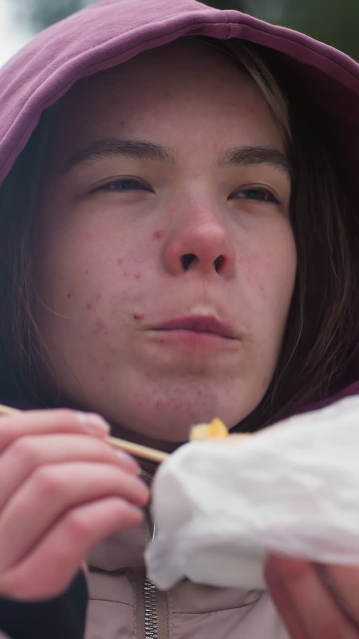 primo piano di una giovane donna in giacca d'inverno che mangia mais, impacchettata nel freddo, godendo di caldo cibo di strada, giacca con cappuccio, spuntino avvolto, tenendo il cibo con un tovagliolo, alberi sfocati e sfondo invernale