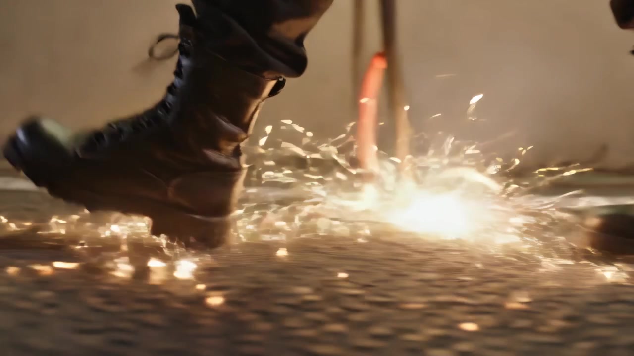 Gehende schwarze Kampfstiefel treten über Asphalt, ein Stiefel passiert ein oranges Rad, Funken sprühend.