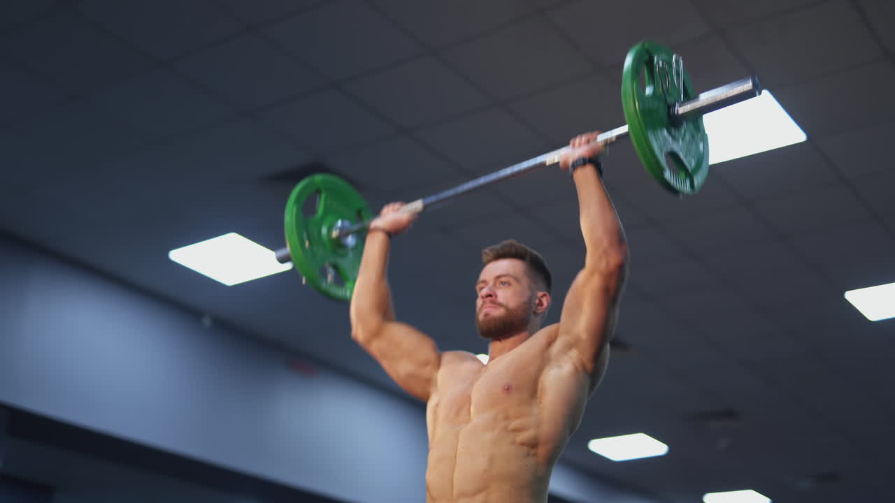 Bodybuilder raising barbell over his head. Young shirtless sportsman exercises his maintain muscle in gym. The active man lifting a barbell. Handsome athlete doing weight lifting.