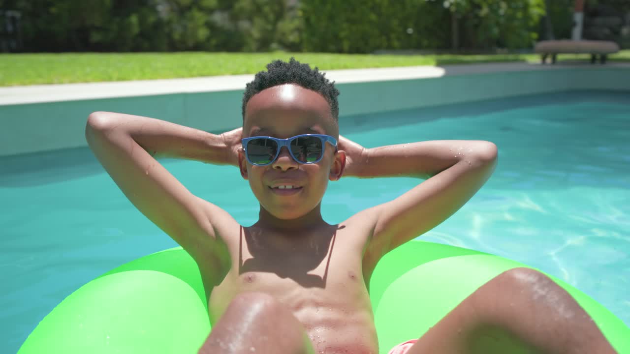 African American youth reclining on green tube in backyard pool squinting from sun relaxing