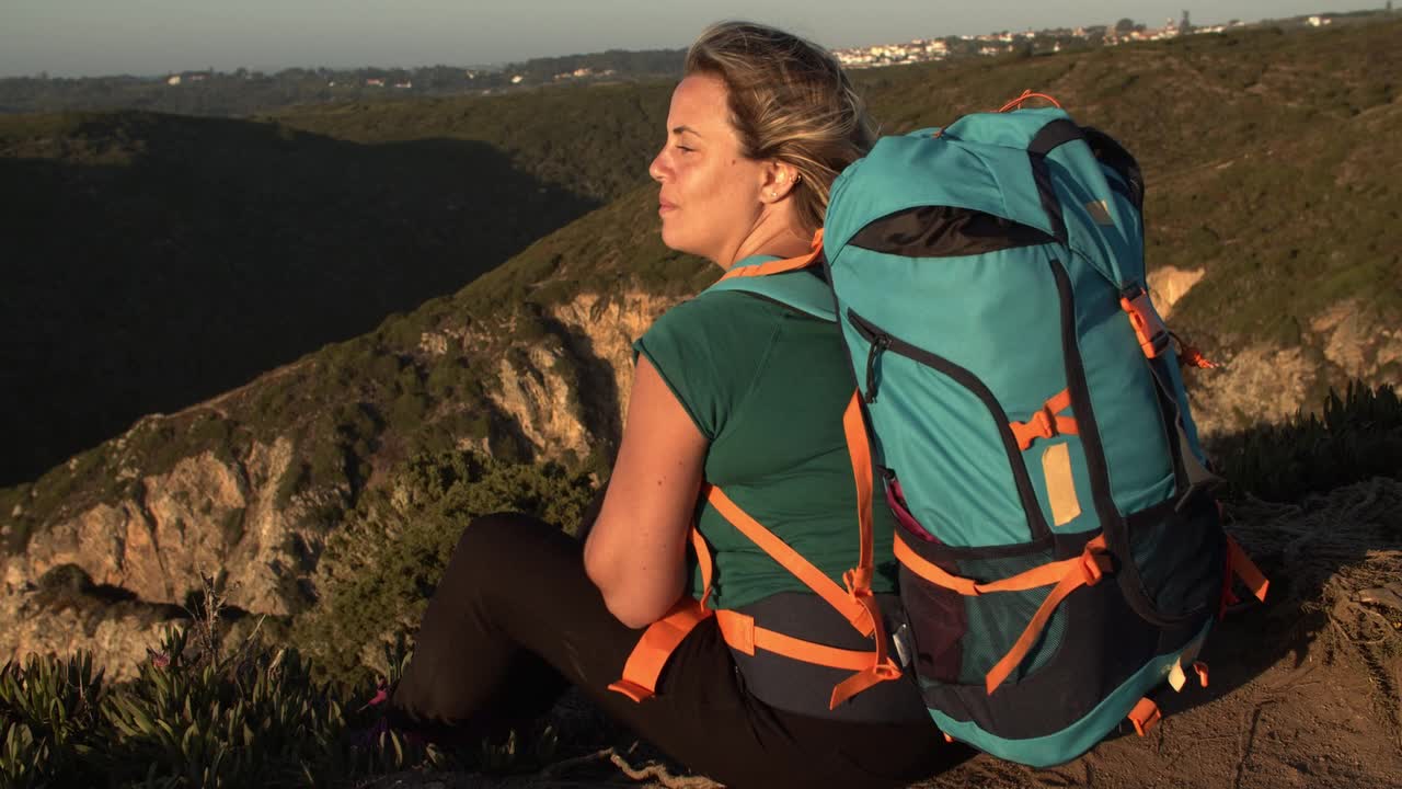 feliz mujer mochilera sentada en un acantilado, mirando el atardecer