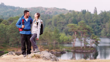 Una-Pareja-De-Adultos-Jóvenes-De-Pie-A-La-Izquierda-Del-Disparo-Contra-Una-Vista-Al-Lago,-Riendo-A-La-Cámara,-Lake-District,-Reino-Unido