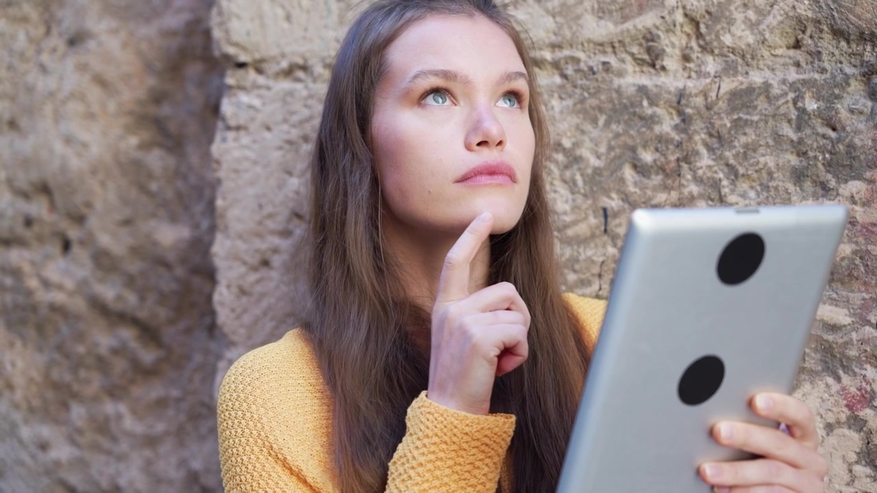 A young woman is happily engaged with her tablet while sitting outdoors against a stone wall