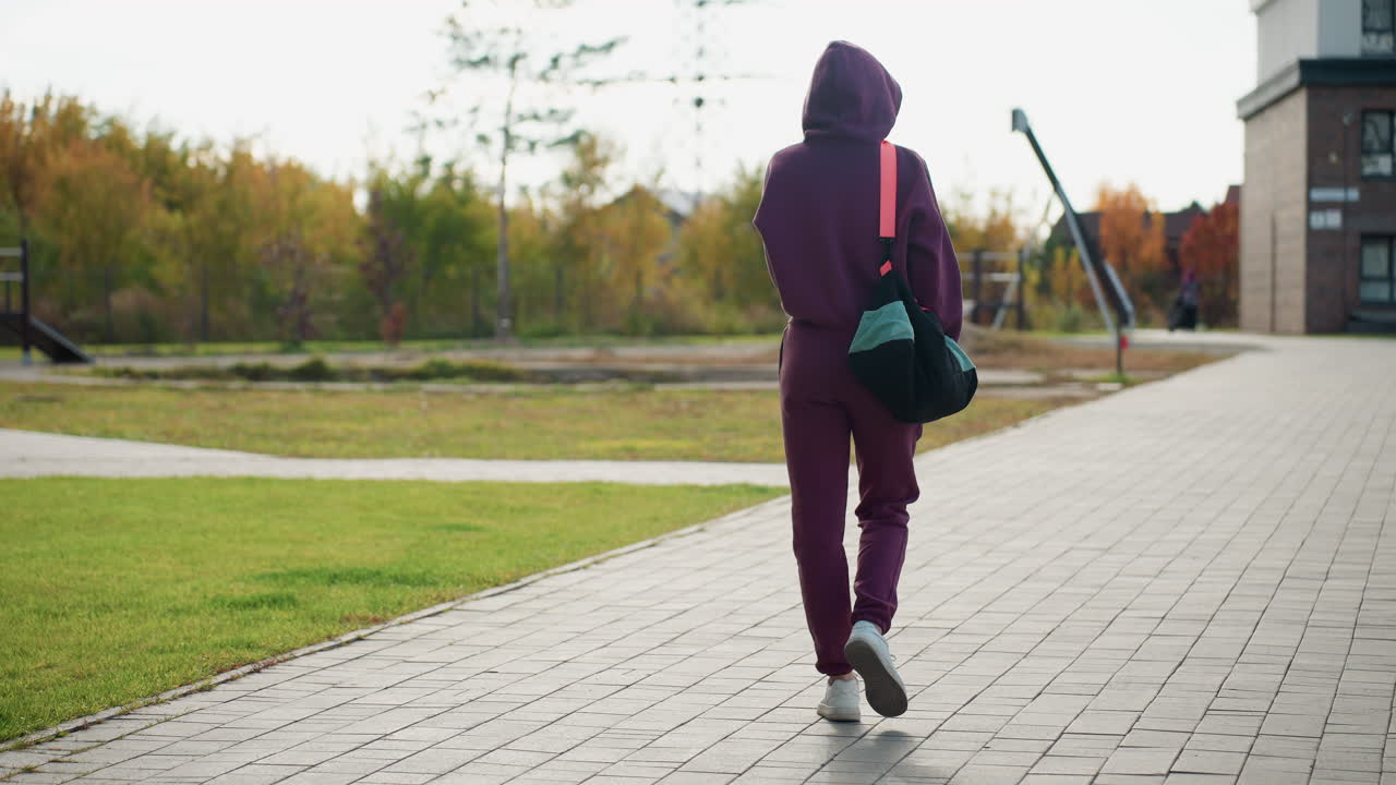Back view of self driven female carrying sport bag walking towards modern building along gray tiled pavement beside green lawn under bright autumn sky with autumn foliage