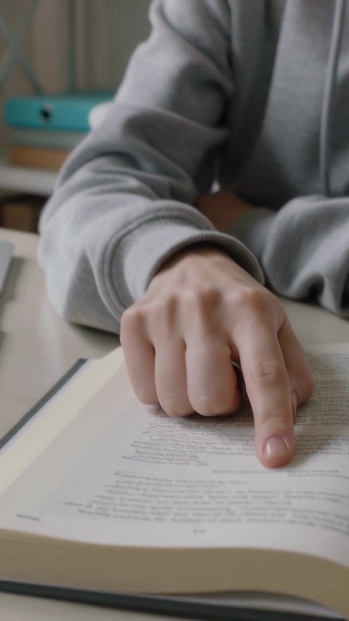 Video verticale: Donna a destra con felpa grigia con cappuccio che guarda il laptop e ripassa un libro alla scrivania studiando.