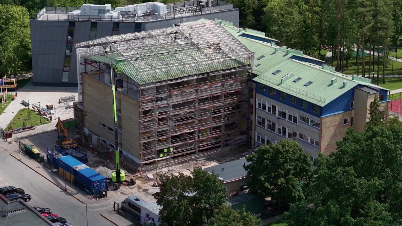 Ilmakuva rakenteilla olevasta rakennuksesta, jossa on laajat rakennustelineet, koneisto ja nosturi toiminnassa aurinkoisena päivänä, korostaen dynaamista kaupunkikehityksen edistymistä.