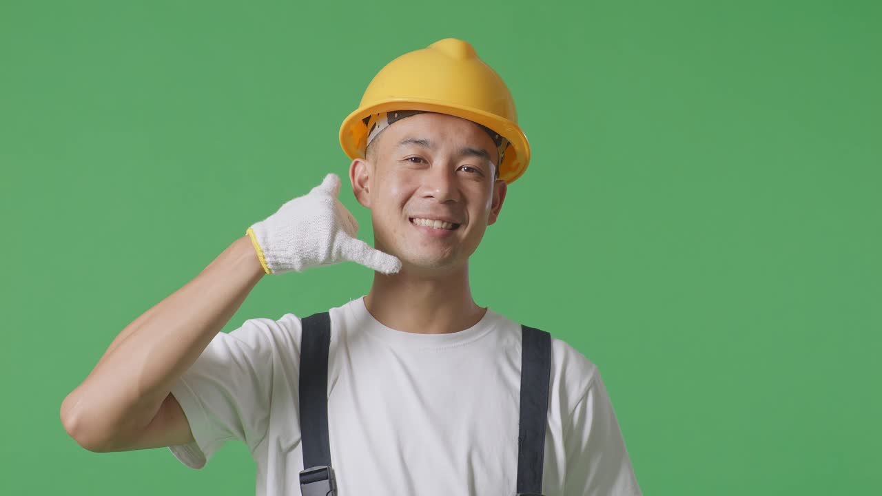 Close Up Of Asian Man Painter Wearing Safety Helmet Smiling And Making Call Me Gesture To Camera While Standing In The Green Screen Background Studio