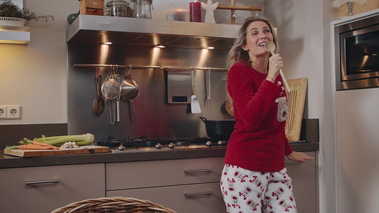Una giovane donna che canta e balla con un maglione natalizio festivo nella sua cucina, mentre prepara un pasto e si diverte ascoltando musica.